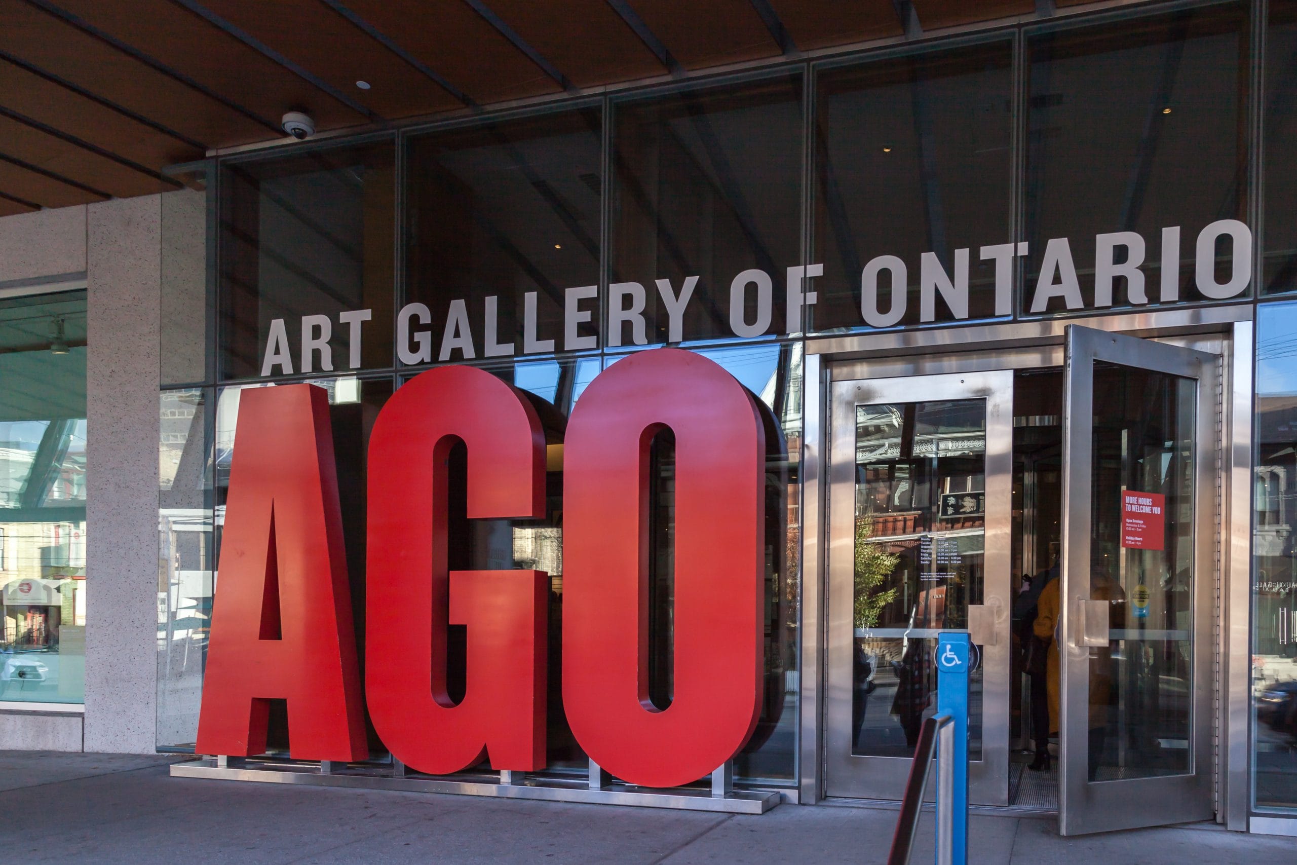 Large red letters spelling AGO stand in front of the glass doors at the Art Gallery of Ontario, with the gallery name above. Reflections of Ontario buildings are visible in the glass.