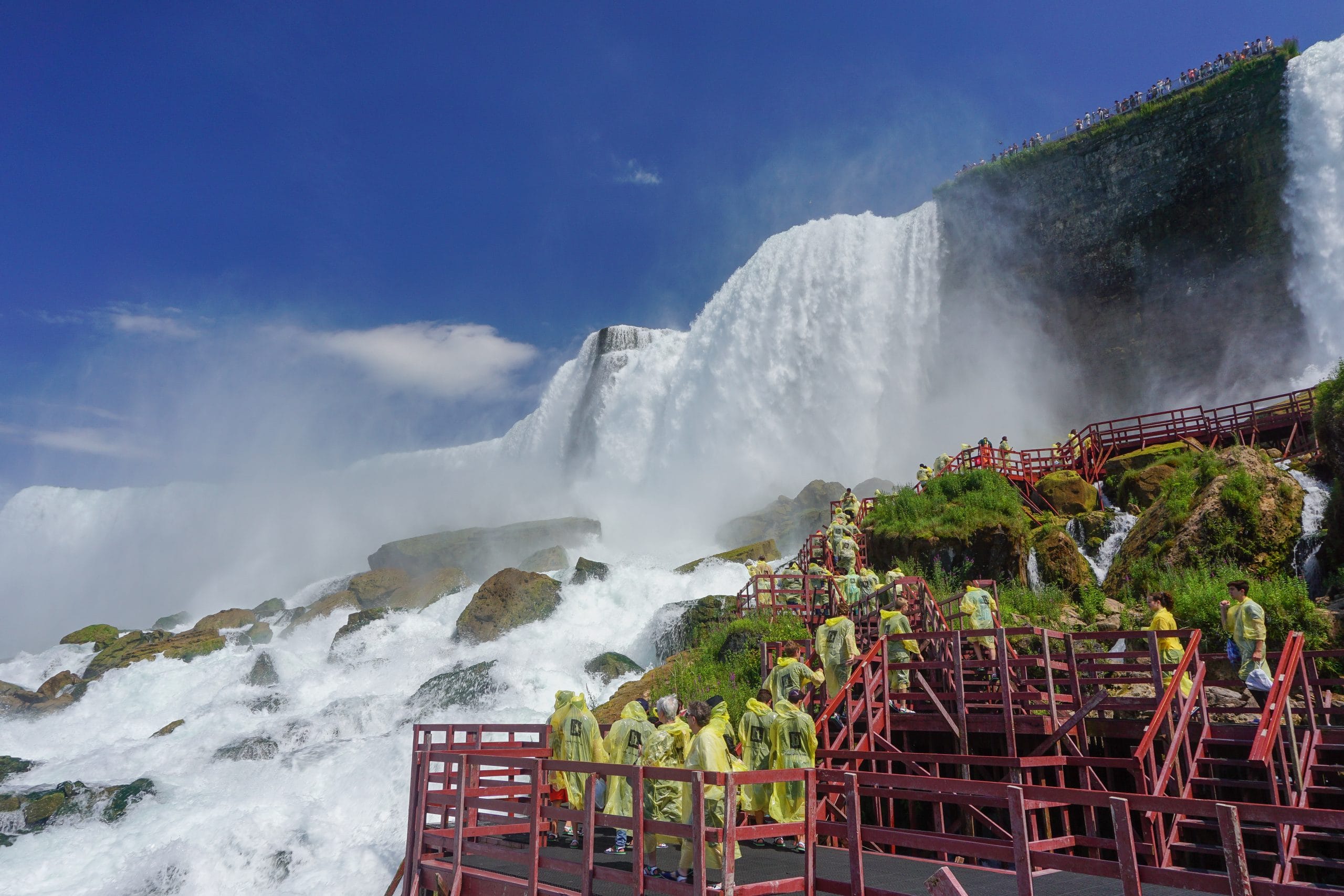 Tourists in yellow rain ponchos walk on red wooden platforms near the powerful Cave of the Winds waterfall, with mist rising and lush green rocks around, under a bright blue sky.