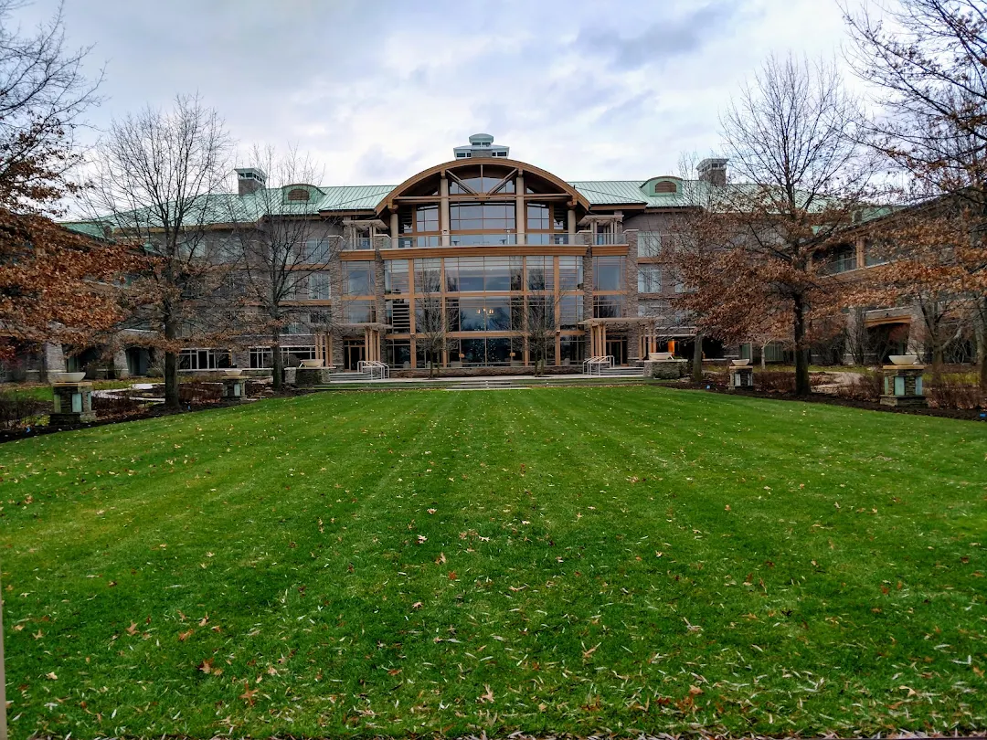The Lodge at Turning Stone, a luxury resort with a glass and wood facade, stands across a green lawn, surrounded by leafless trees beneath a cloudy sky.