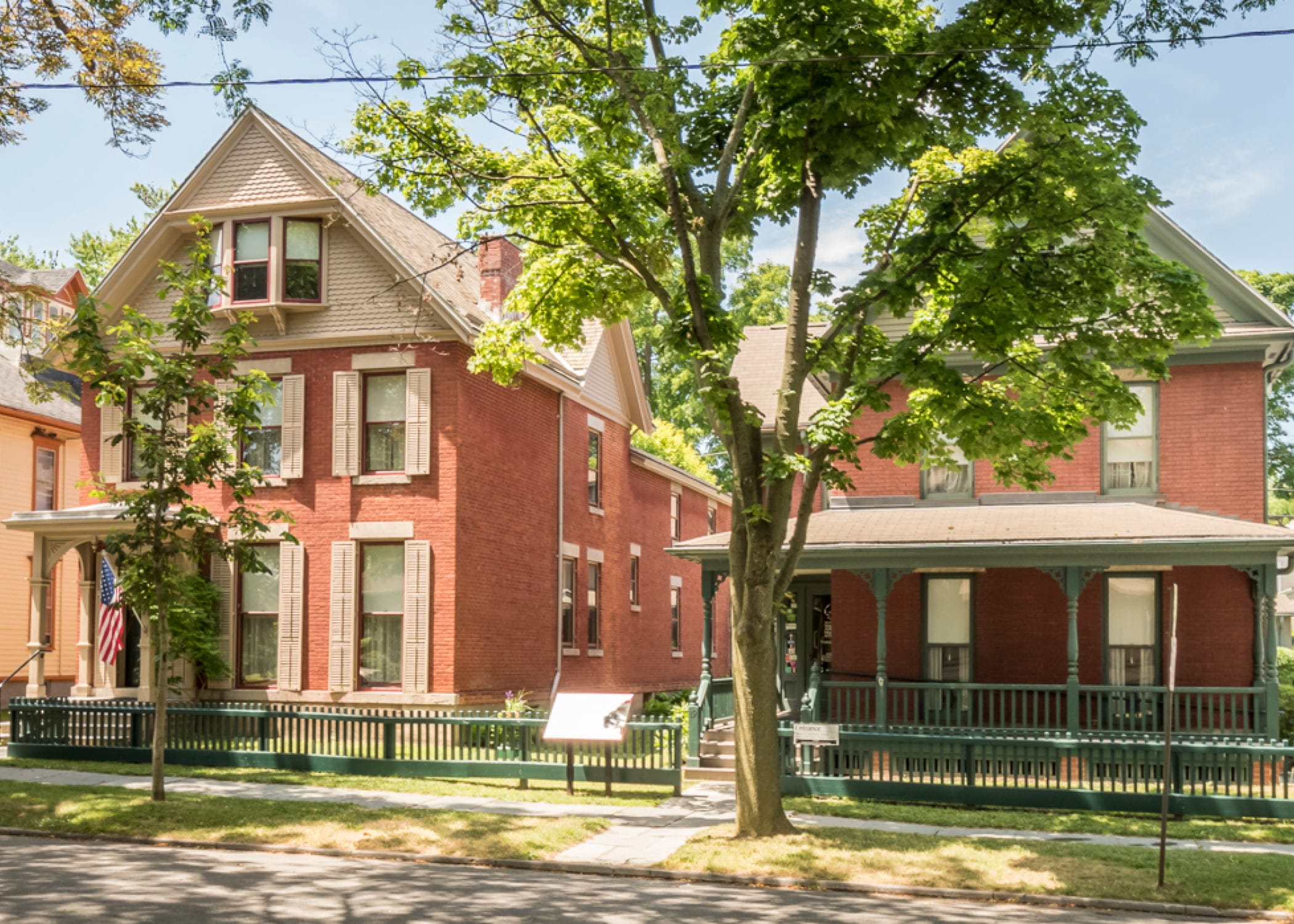 A sunny street view of two adjacent red-brick houses with green lawns and trees resembles the charming surroundings near the Susan B. Anthony Museum. One house has a peaked roof, while the other features a porch with a green railing. An American flag proudly waves in front of one house. A sunny street view of two adjacent red-brick houses with green lawns and trees resembles the charming surroundings near the Susan B. Anthony Museum. One house has a peaked roof, while the other features a porch with a green railing. An American flag proudly waves in front of one house.