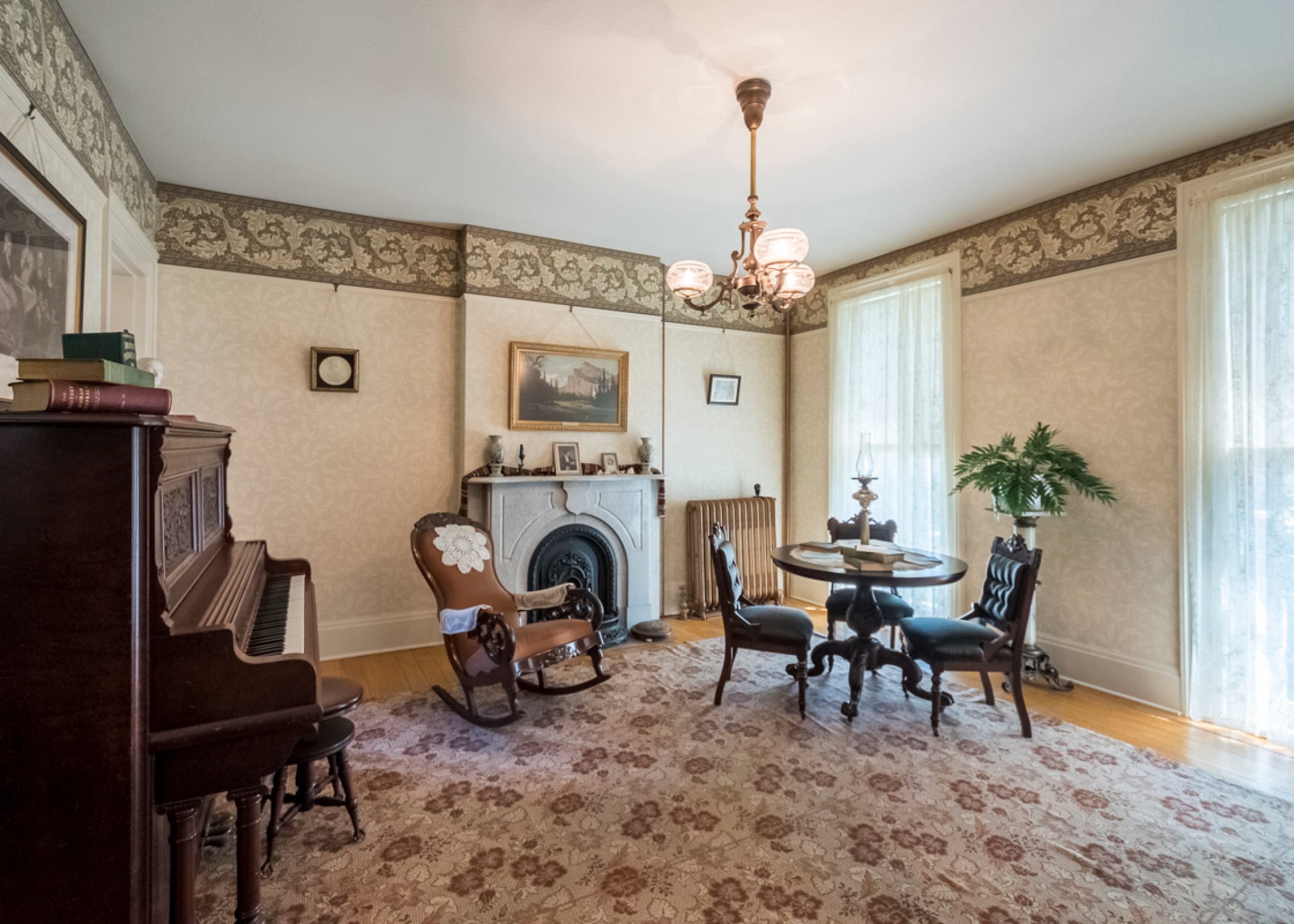 A vintage living room reminiscent of the Susan B. Anthony Museum features a fireplace, rocking chair, and piano. Patterned wallpaper adorns the walls, while large windows with sheer curtains welcome natural light. The ambiance is completed by an ornate chandelier and a round table with chairs. A vintage living room reminiscent of the Susan B. Anthony Museum features a fireplace, rocking chair, and piano. Patterned wallpaper adorns the walls, while large windows with sheer curtains welcome natural light. The ambiance is completed by an ornate chandelier and a round table with chairs.