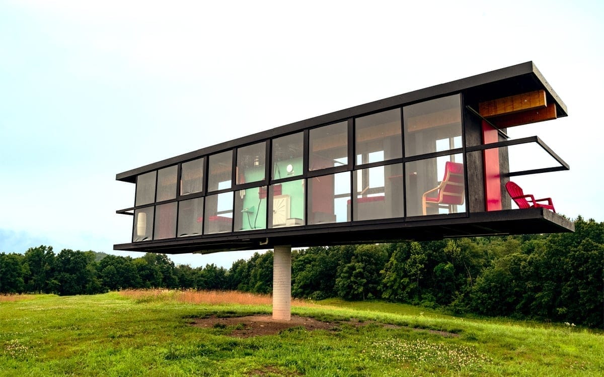 A modern, glass-walled house in Columbia County is elegantly elevated on a central pedestal, standing proudly in an open grassy field with trees in the background. Close to Art Omi, this rectangular structure features large windows that showcase vibrant furniture and intricate interior details. A modern, glass-walled house in Columbia County is elegantly elevated on a central pedestal, standing proudly in an open grassy field with trees in the background. Close to Art Omi, this rectangular structure features large windows that showcase vibrant furniture and intricate interior details.