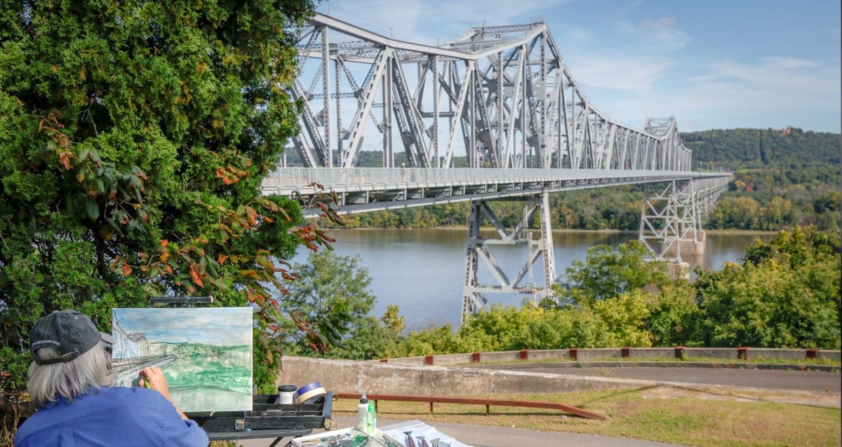 A person with gray hair paints a landscape scene featuring a large steel bridge over a river, reminiscent of the Hudson River Skywalk. Lush green trees surround the area, and hills are visible in the background under a clear blue sky.