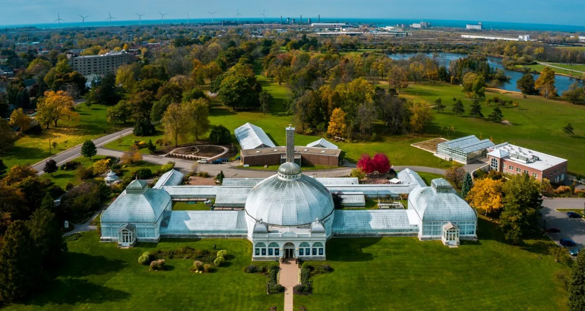 From an aerial view, a large historic greenhouse with a glass dome sits amidst lush green lawns and autumn trees. In the background, wind turbines and the cityscape peek through under a clear blue sky, reminiscent of the Buffalo and Erie Canals blend of nature and industry.