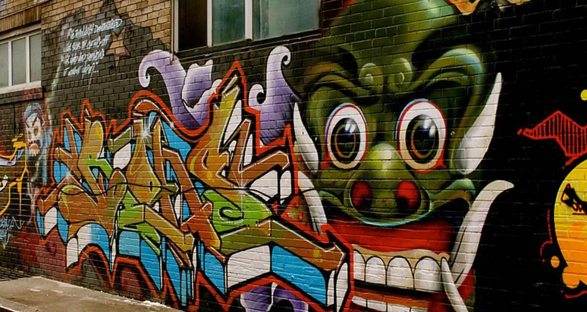 A vibrant graffiti mural on a brick wall features colorful abstract shapes and a large, detailed green face with exaggerated features, including wide eyes and a big grin.