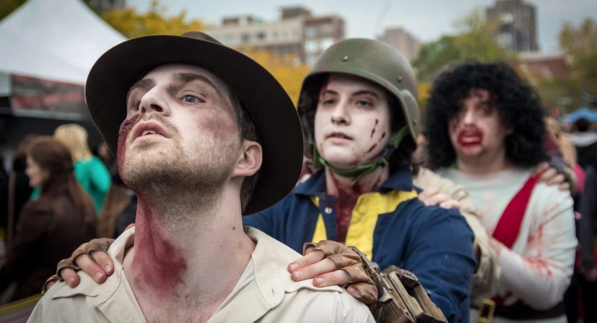 Three people in costume, wearing makeup to appear as zombies, march in a line at the Montreal Zombie Walk. The first person wears a hat, the second a helmet, and the third has long dark hair. Buildings and autumn trees create an eerie backdrop in the crowded outdoor area.