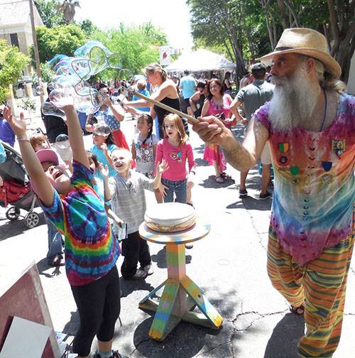 A bearded man in colorful clothes makes bubbles with a wand at the Park Avenue Summer Arts Festival. Children eagerly reach for the bubbles, smiling and enjoying the sunny day. Trees and vibrant booths are visible in the background, adding to the festivals charm.