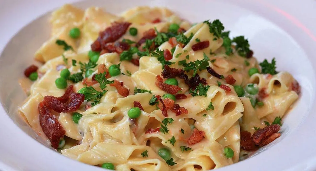 A plate of creamy pasta garnished with green peas, crispy bacon pieces, and chopped parsley. The pasta appears to be coated in a rich sauce, adding a savory and colorful touch to the dish.