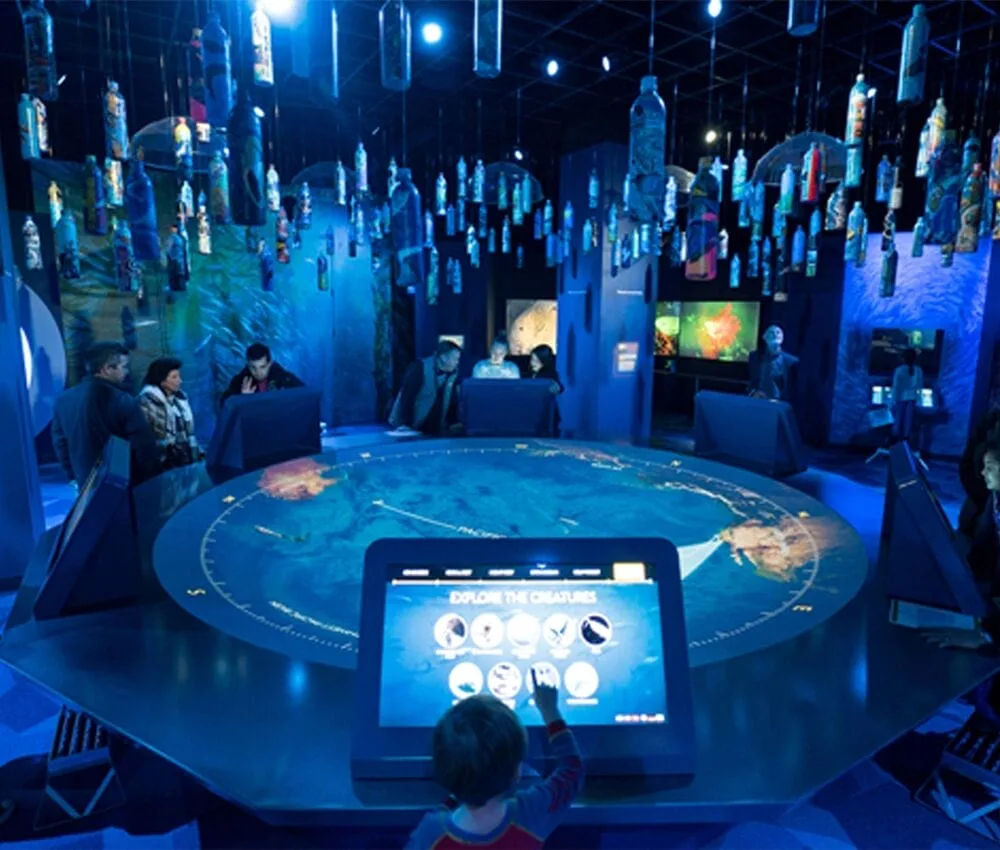 A child interacts with a digital display in a dimly lit, futuristic National Geographic exhibit. People sit around a large circular table with a world map. Numerous hanging bottles are illuminated above, creating a captivating atmosphere.