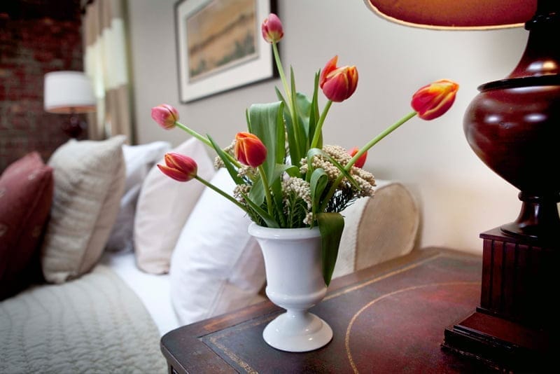 A white vase filled with vibrant red and orange tulips sits on a wooden nightstand at the Morgan State House Inn. The background features a cozy bed with multiple pillows and a lamp, creating a warm, inviting atmosphere. A white vase filled with vibrant red and orange tulips sits on a wooden nightstand at the Morgan State House Inn. The background features a cozy bed with multiple pillows and a lamp, creating a warm, inviting atmosphere.