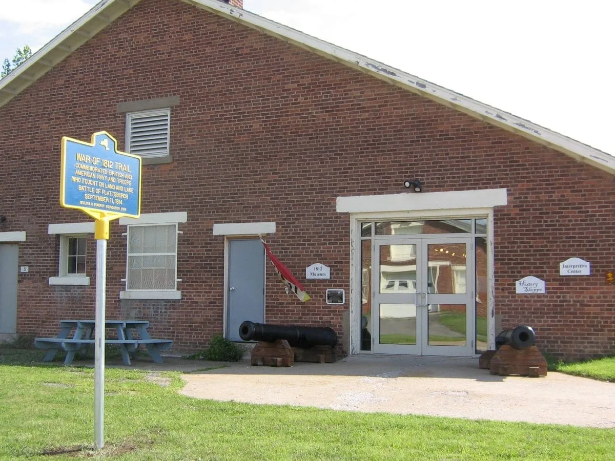 A red brick building showcases historical cannons outside and features a sign, War of 1812 Trail. A blue picnic table sits on the grassy area in front. The building, which resembles a museum, also has a library sign above its glass doors.
