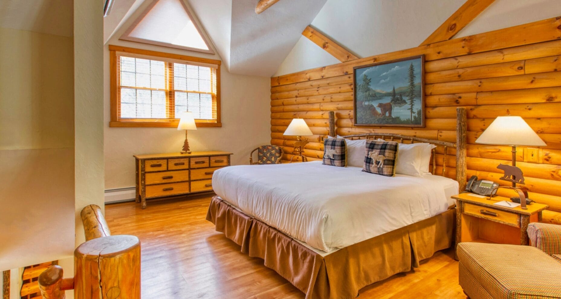 A cozy log cabin bedroom at the Catskills Resort & Spa features a large bed, plaid pillows, and a wooden nightstand. Soft lighting from two lamps illuminates the warm wooden walls, creating an inviting atmosphere. A scenic painting above the bed adds a rustic touch.