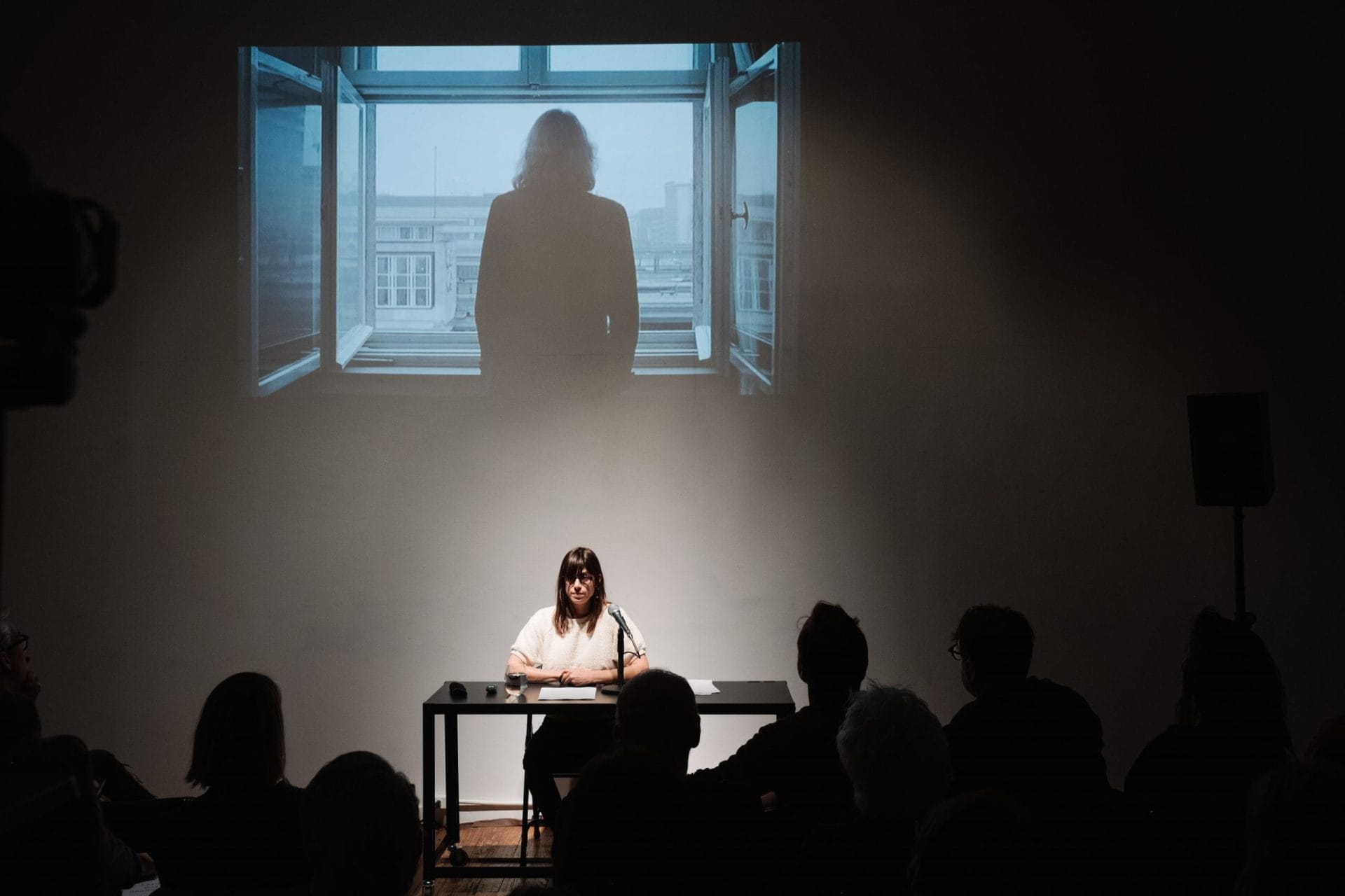 A woman sits at a table speaking to an audience in a dimly lit room. Above her, a projected virtual image shows a person standing by an open window, looking out. The setting suggests a presentation or lecture environment.