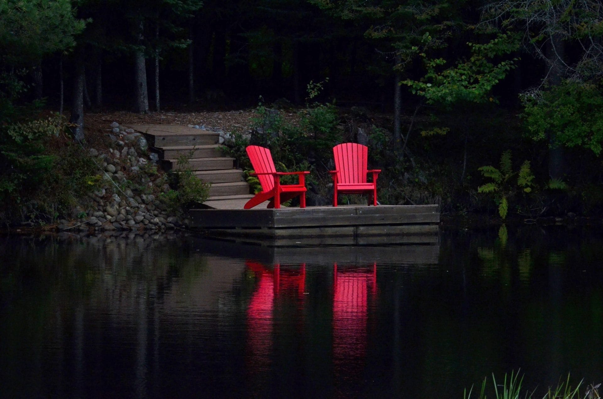 Two red Adirondack chairs are on a wooden dock by a calm lake, surrounded by lush green trees. Like an exquisite piece at a sidewalk art show, the reflection of the chairs shimmers on the waters surface, painting an idyllic and tranquil scene. Two red Adirondack chairs are on a wooden dock by a calm lake, surrounded by lush green trees. Like an exquisite piece at a sidewalk art show, the reflection of the chairs shimmers on the waters surface, painting an idyllic and tranquil scene.