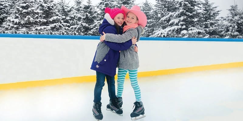 Two children in colorful winter clothes and hats hug while ice skating outdoors at Rocking Horse Ranch Resort. Snow-covered trees line the background, creating a festive winter scene. Two children in colorful winter clothes and hats hug while ice skating outdoors at Rocking Horse Ranch Resort. Snow-covered trees line the background, creating a festive winter scene.