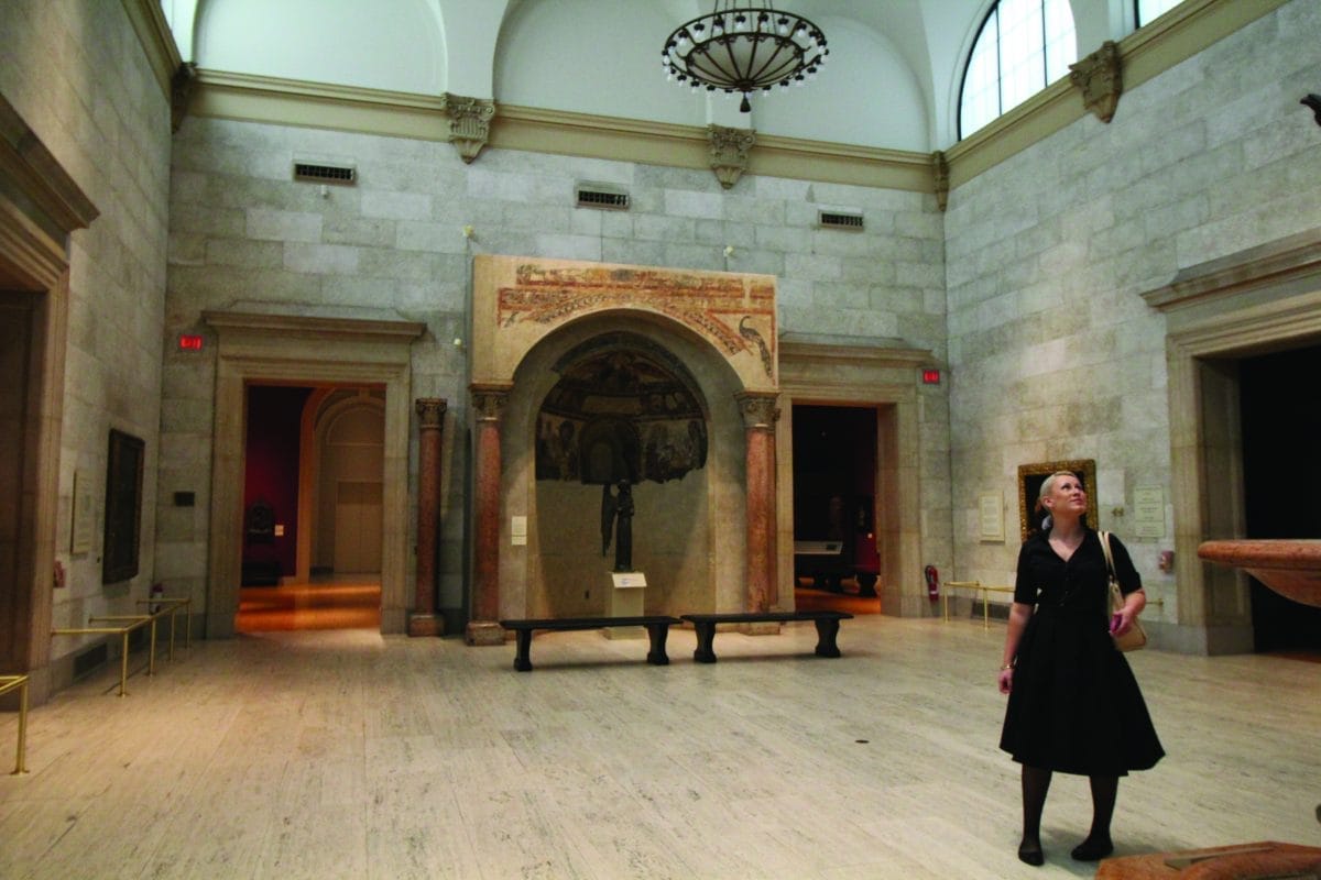 A woman in a black dress stands in a spacious memorial art gallery with high ceilings. She gazes upward, surrounded by paintings and sculptures. A large archway painting adorns the central wall. The room is softly lit, creating a serene atmosphere.