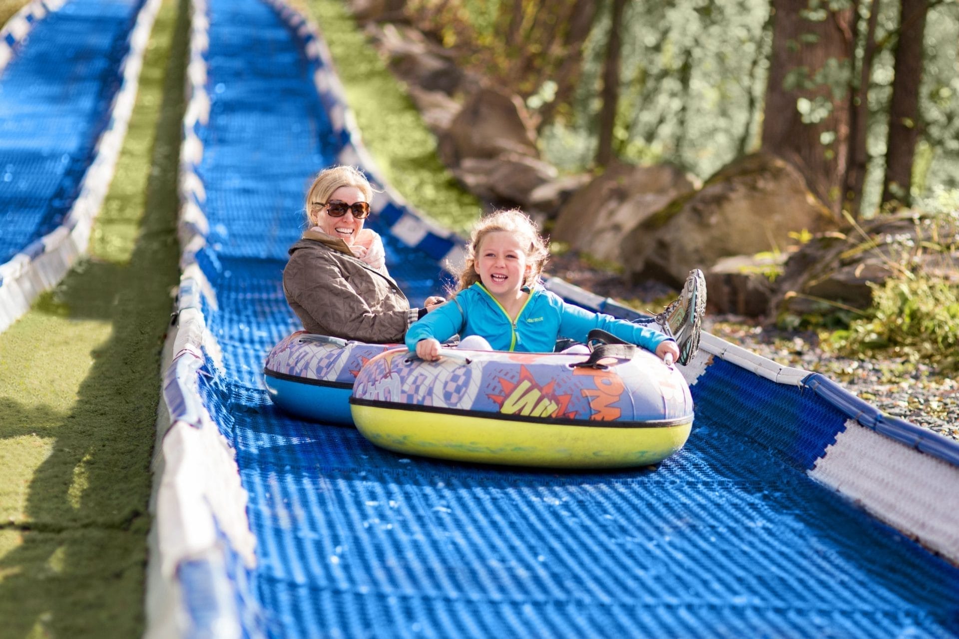 A woman and a child are smiling as they slide down an outdoor incline in colorful inflatable tubes at Rocking Horse Ranch Resort. The slide is blue and green, surrounded by trees and rocks. Its a sunny day, adding a cheerful atmosphere to the activity. A woman and a child are smiling as they slide down an outdoor incline in colorful inflatable tubes at Rocking Horse Ranch Resort. The slide is blue and green, surrounded by trees and rocks. Its a sunny day, adding a cheerful atmosphere to the activity.