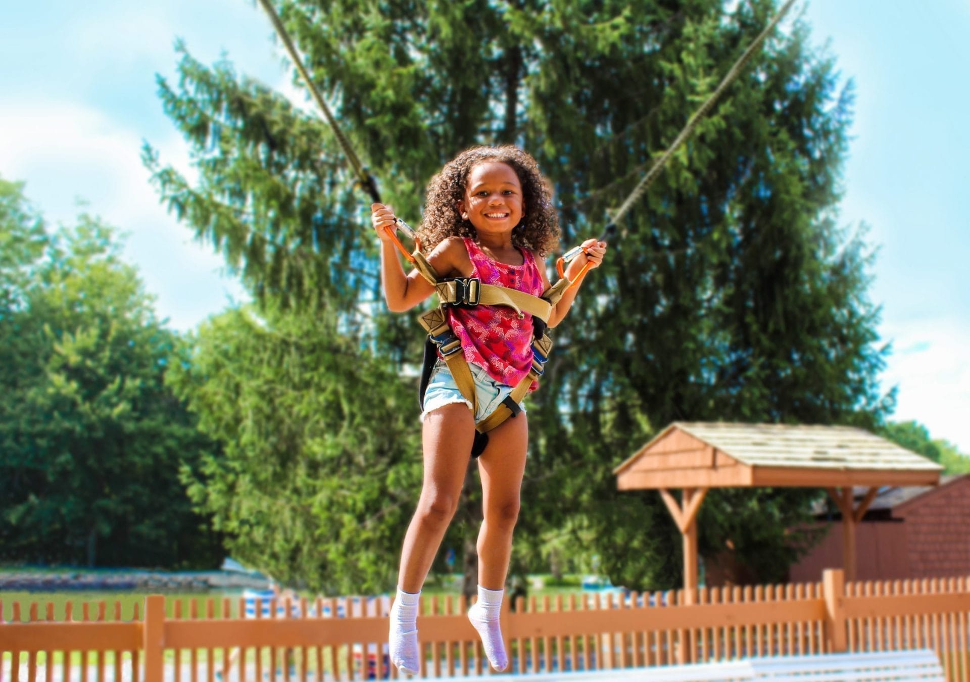 A young girl with curly hair is joyfully jumping on a trampoline, wearing safety harnesses at Rocking Horse Ranch Resort. She is outdoors with trees and a fence in the background, while the sunlight illuminates the scene. A young girl with curly hair is joyfully jumping on a trampoline, wearing safety harnesses at Rocking Horse Ranch Resort. She is outdoors with trees and a fence in the background, while the sunlight illuminates the scene.
