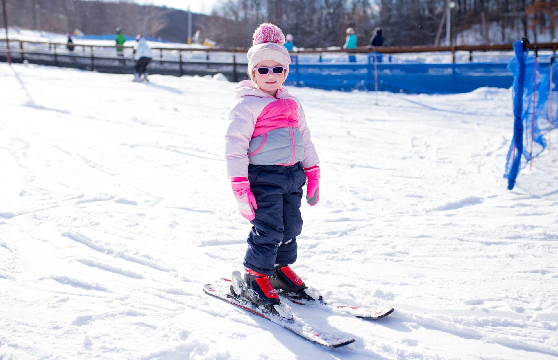 A young child in a pink jacket, matching hat, and sunglasses stands confidently on skis at Rocking Horse Ranch Resort. People ski in the background under a clear sky, surrounded by bright snow. A young child in a pink jacket, matching hat, and sunglasses stands confidently on skis at Rocking Horse Ranch Resort. People ski in the background under a clear sky, surrounded by bright snow.