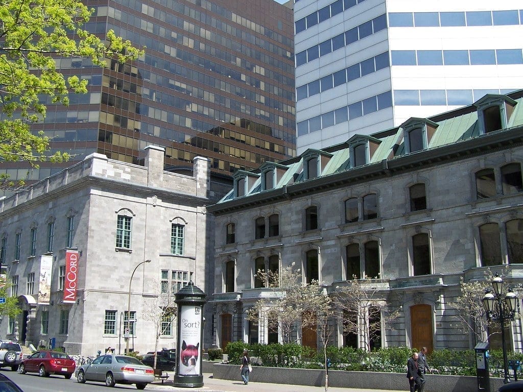 The street view of the McCord Museum in Montreal showcases its gray stone architecture with a distinctive green roof. People amble along the sidewalk, framed by modern high-rises and trees with sparse leaves lining the bustling street.
