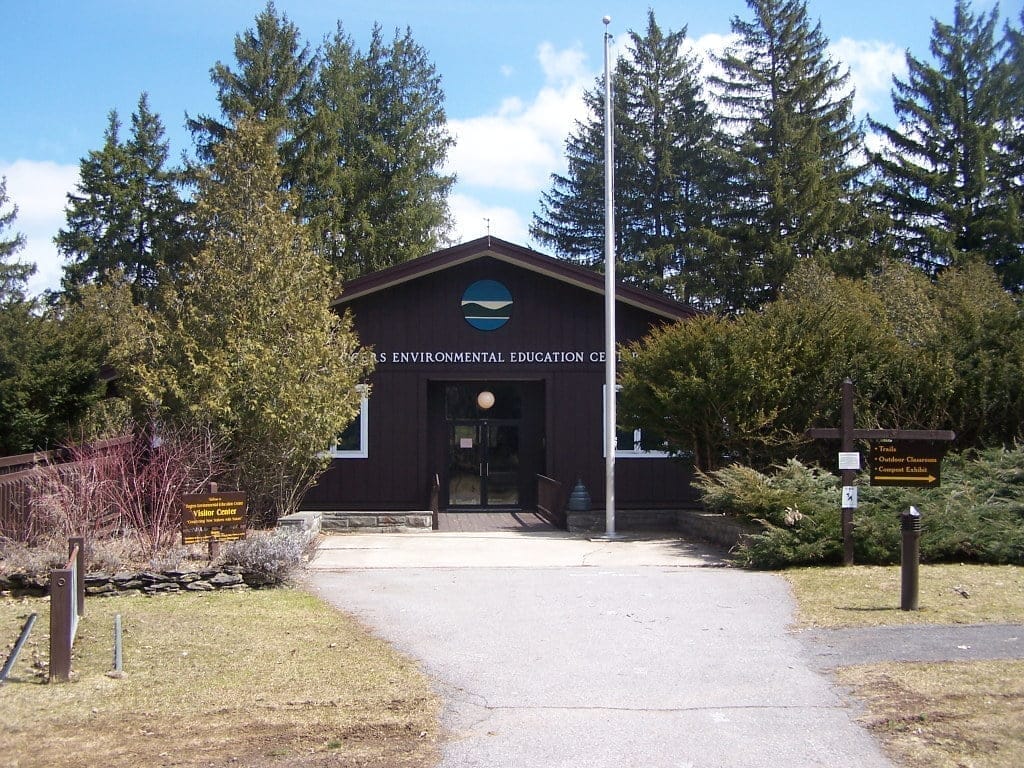 A brown wooden building labeled Environmental Education Center is surrounded by tall evergreen trees. A clear pathway leads to the entrance, and signs provide directions. The sky is blue, suggesting a sunny day. A brown wooden building labeled Environmental Education Center is surrounded by tall evergreen trees. A clear pathway leads to the entrance, and signs provide directions. The sky is blue, suggesting a sunny day.