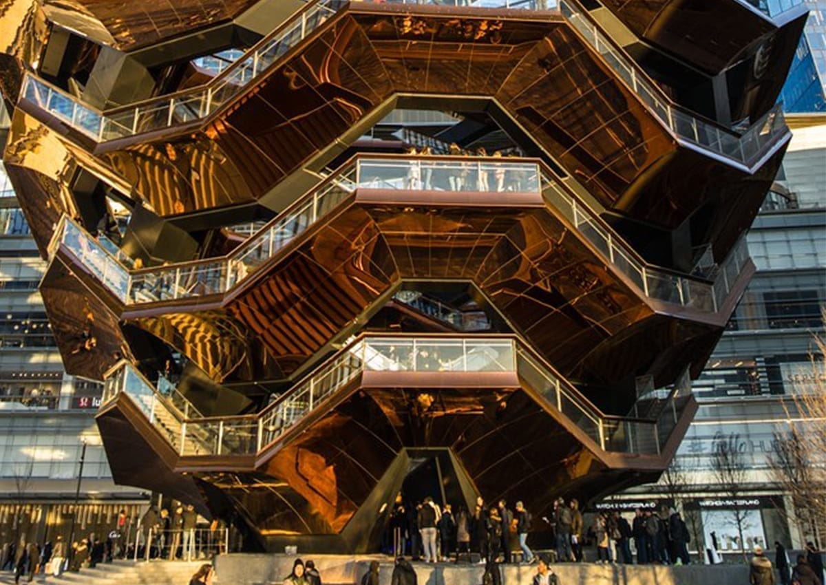 A large, honeycomb-like metal and glass structure with multiple levels and staircases, the Vessel at Hudson Yards stands majestic. People are walking and standing on various levels, surrounded by modern buildings and bathed in warm sunlight.