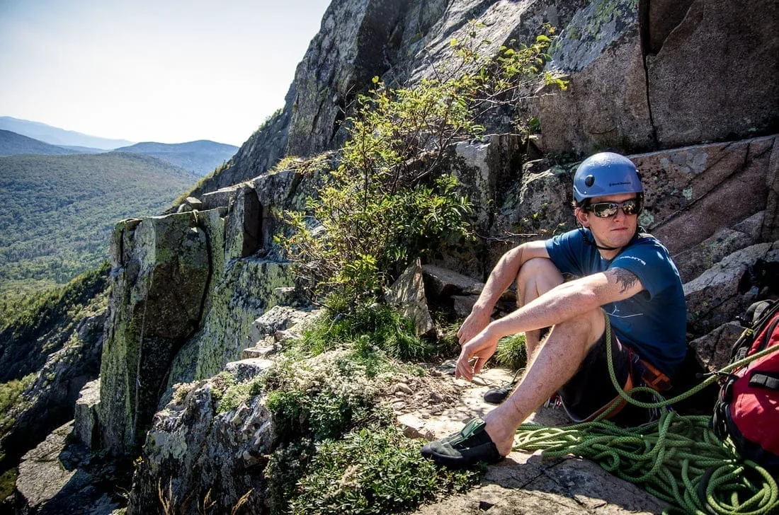 A person wearing a helmet and sunglasses takes a break while rock climbing, seated on a rocky ledge with a coiled rope. As part of Vermont Adventure Tours, the backdrop offers an outdoor attraction: scenic mountain views and lush greenery under the clear blue sky.