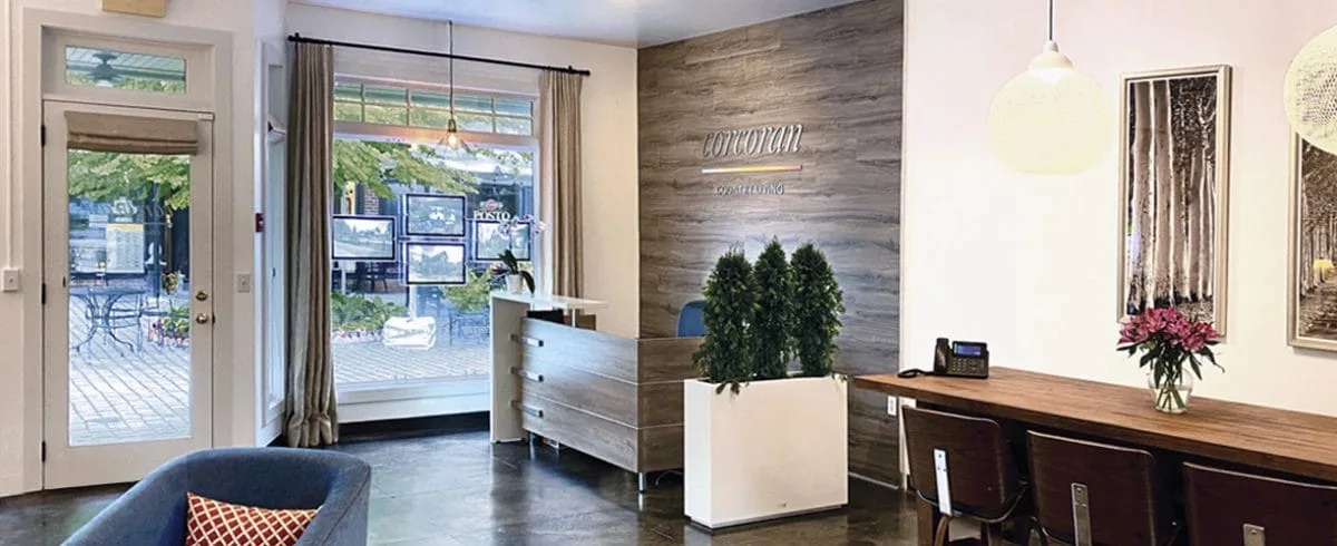 A modern office reception area with a wooden desk, chairs, and wall art exudes the charm of Corcoran Country Living. A window overlooks a patio with trees. Warm lighting highlights a plant and flower vase, while a sign on the wall reads FUNOVA.