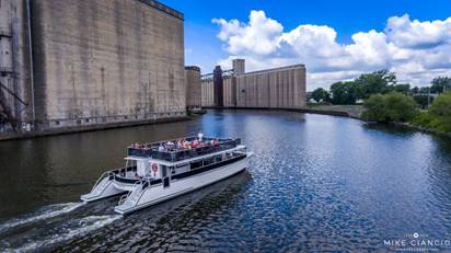 A sightseeing boat from Buffalo River History Tours glides along the river, navigating between large industrial buildings. The sky is blue with scattered clouds, and lush vegetation lines the riverbank in the background.