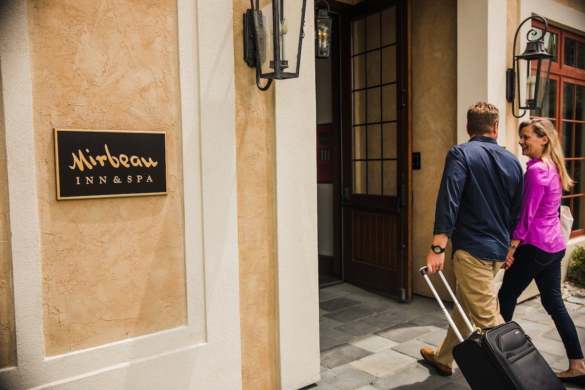 A man and woman are entering the Mirbeau Inn & Spa in Rhinebeck, the man pulling a suitcase. The building features a beige exterior with a welcoming sign beside the door.