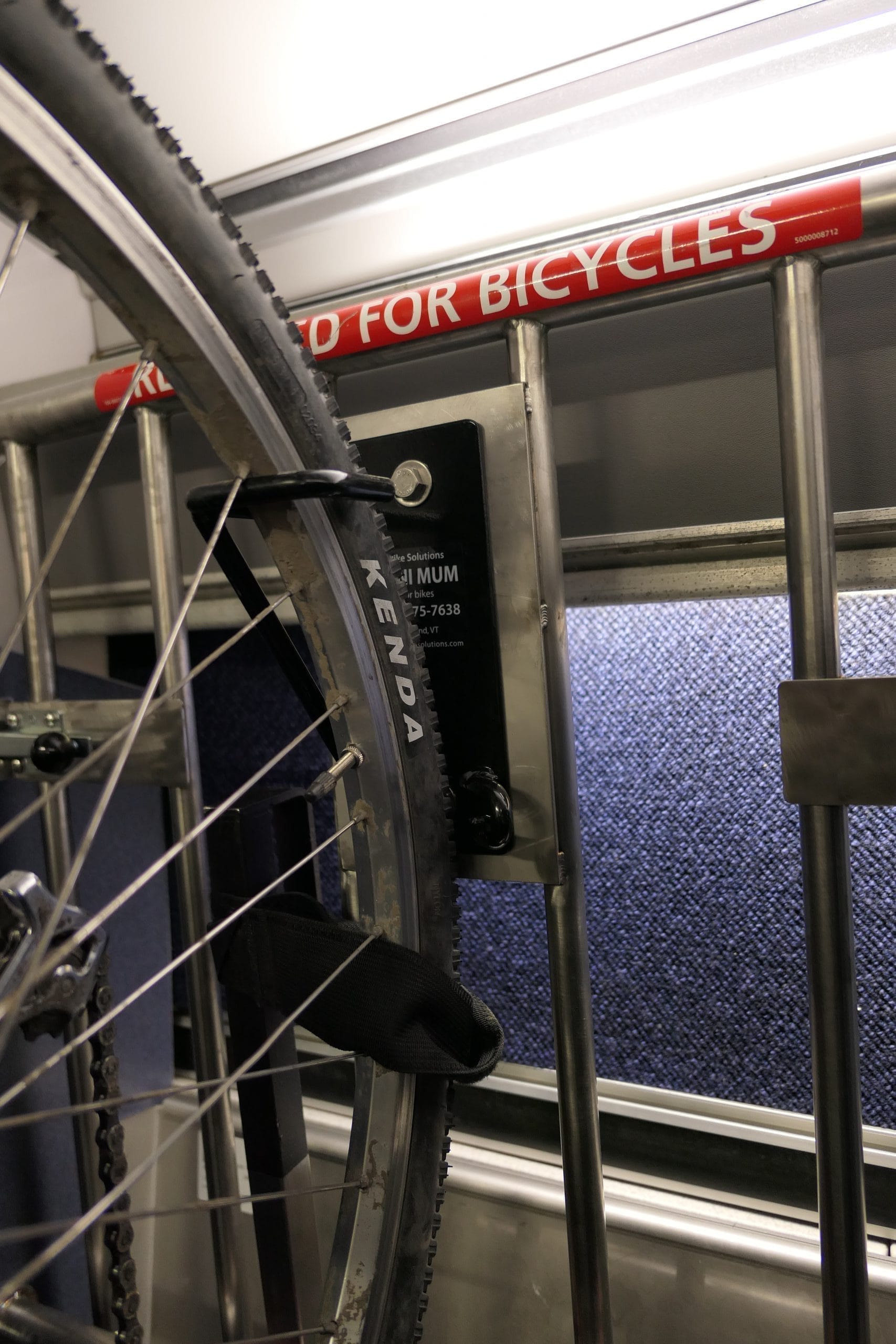 A bike is secured on a train with a strap around the front wheel. A sign above reads PRIORITY SPACE FOR BICYCLES. The tire is labeled KENDA. In the background, a blue textured panel adds contrast to the scene. A bike is secured on a train with a strap around the front wheel. A sign above reads PRIORITY SPACE FOR BICYCLES. The tire is labeled KENDA. In the background, a blue textured panel adds contrast to the scene.