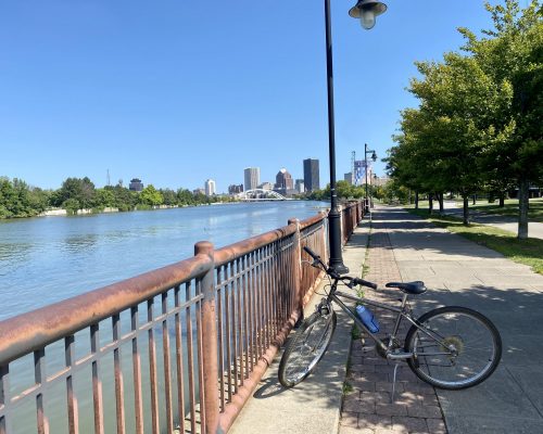 A bike rests against a riverside railing on a sunny day. The city skyline with its tall buildings and bridge stretches across the river. A tree-lined path follows the railing under the clear blue sky, offering a perfect spot for a leisurely ride or stroll. A bike rests against a riverside railing on a sunny day. The city skyline with its tall buildings and bridge stretches across the river. A tree-lined path follows the railing under the clear blue sky, offering a perfect spot for a leisurely ride or stroll.