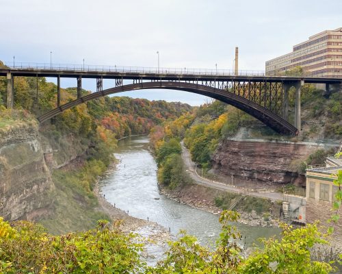 A wide shot of a river cutting through a gorge adorned with lush autumn-colored foliage. A large arch bridge spans the gorge, where bikers occasionally glide by. On the right, a tall building stands against the overcast sky. A wide shot of a river cutting through a gorge adorned with lush autumn-colored foliage. A large arch bridge spans the gorge, where bikers occasionally glide by. On the right, a tall building stands against the overcast sky.