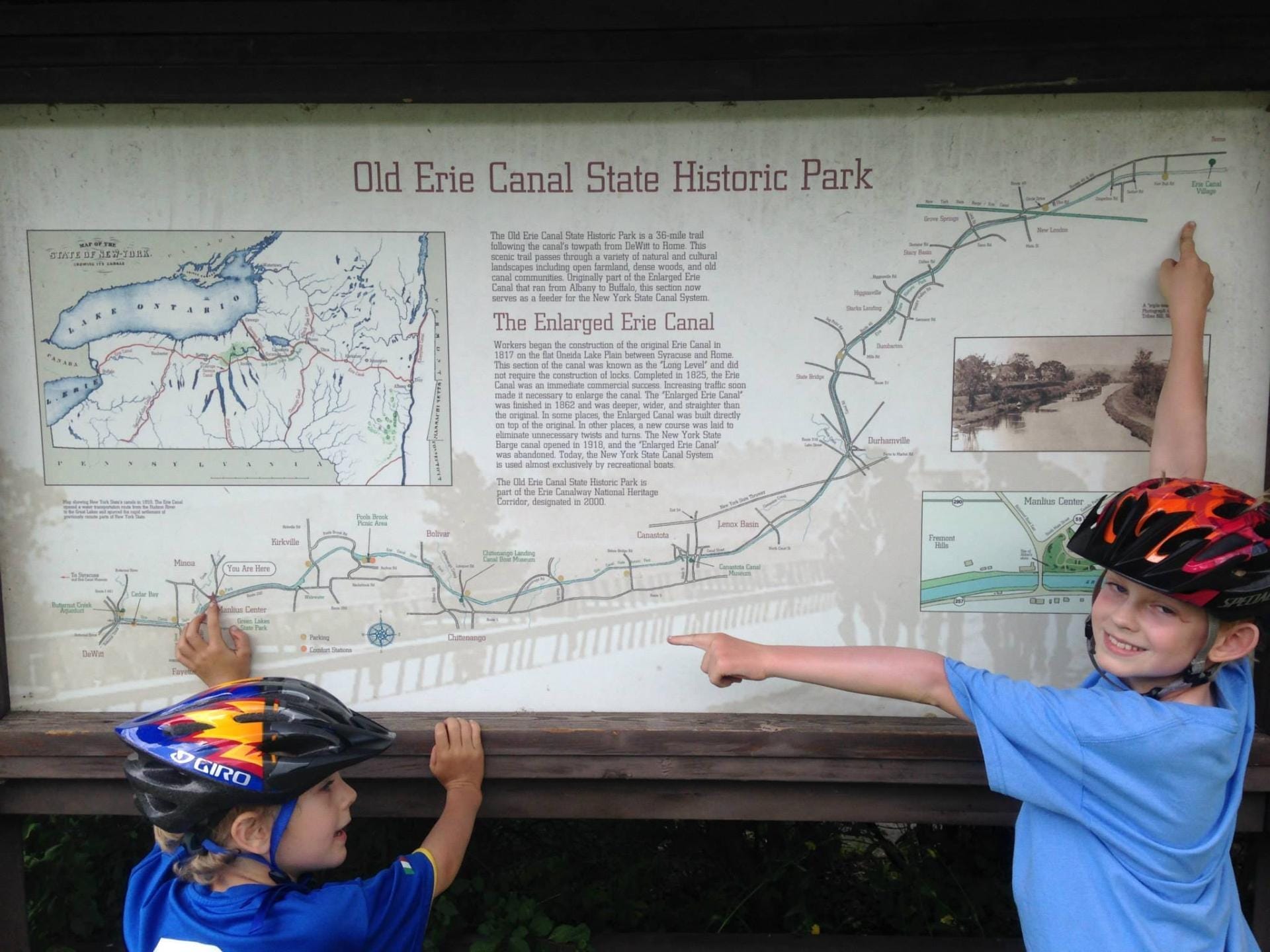 Two children in helmets are pointing at a large informational sign about the Old Erie Canal State Historic Park, likely after an exhilarating bike ride. The sign includes a map, historical text, and images. The children appear engaged and smiling. Two children in helmets are pointing at a large informational sign about the Old Erie Canal State Historic Park, likely after an exhilarating bike ride. The sign includes a map, historical text, and images. The children appear engaged and smiling.