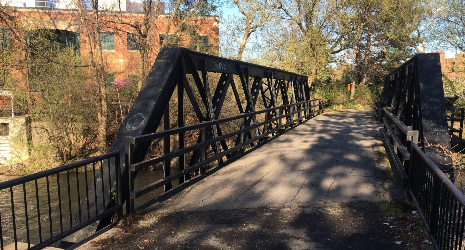 A sleek, black metal bridge provides a tranquil crossing over the calm river, perfect for a leisurely bike ride. Leafy trees embrace the scene, while in the background, a red brick building with green windows adds charm as sunlight and shadows dance along the pathway. A sleek, black metal bridge provides a tranquil crossing over the calm river, perfect for a leisurely bike ride. Leafy trees embrace the scene, while in the background, a red brick building with green windows adds charm as sunlight and shadows dance along the pathway.