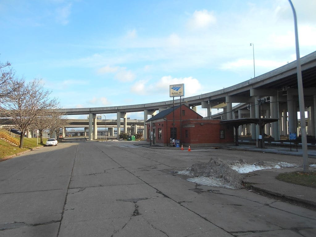 Near the Buffalo-Exchange Street Station, a small red brick building stands close to an overpass with multiple elevated highway ramps. A few traffic cones and a pile of debris are out front, while a white car is parked alongside the cracked road. The sky above is partly cloudy. Near the Buffalo-Exchange Street Station, a small red brick building stands close to an overpass with multiple elevated highway ramps. A few traffic cones and a pile of debris are out front, while a white car is parked alongside the cracked road. The sky above is partly cloudy.