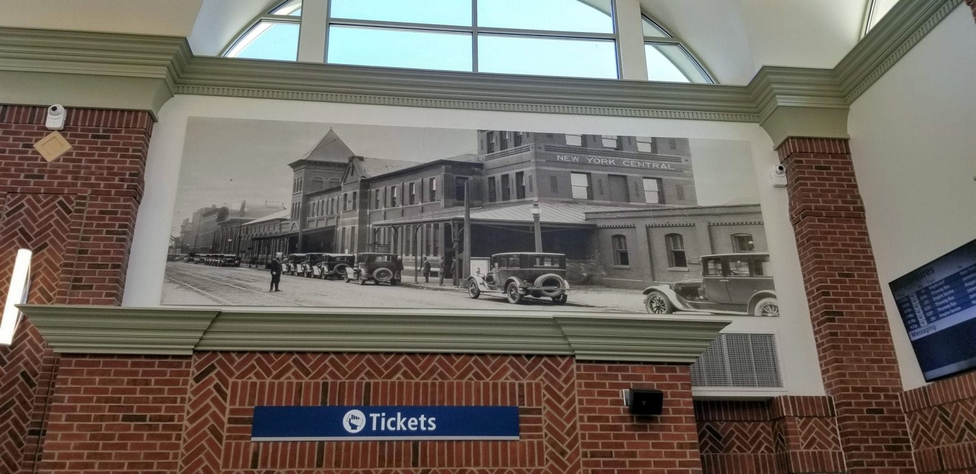 A large black-and-white mural depicting a historic street scene with vintage cars and buildings, including a sign for New York Central, graces the ticketing area of Buffalo-Exchange Street Station, blending the citys rich past with its vibrant present in this modern brick train station. A large black-and-white mural depicting a historic street scene with vintage cars and buildings, including a sign for New York Central, graces the ticketing area of Buffalo-Exchange Street Station, blending the citys rich past with its vibrant present in this modern brick train station.
