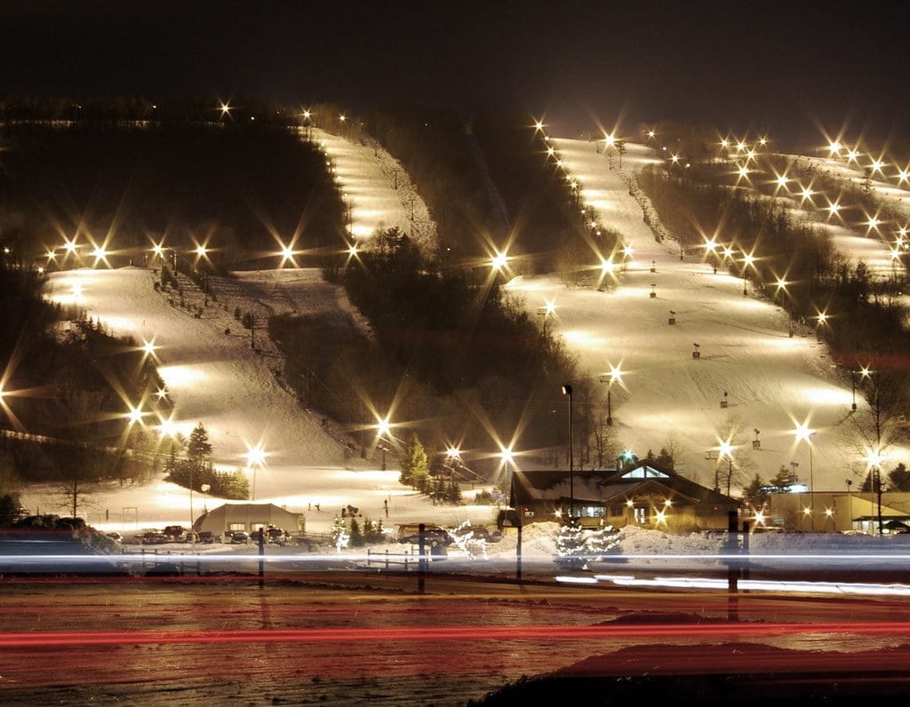 A ski resort at night features illuminated slopes with lights outlining multiple ski trails. The snow-covered landscape glimmers, highlighting the serene winter atmosphere. In the foreground, blurred lights suggest passing vehicles, adding vibrant movement to this ski haven. A ski resort at night features illuminated slopes with lights outlining multiple ski trails. The snow-covered landscape glimmers, highlighting the serene winter atmosphere. In the foreground, blurred lights suggest passing vehicles, adding vibrant movement to this ski haven.