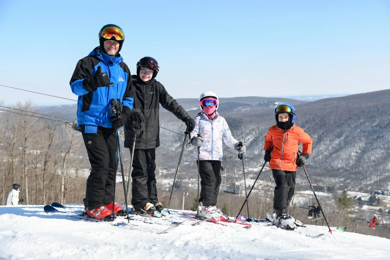 A group of four, including an adult and three children, stand on a snowy ski slope. Clad in ski gear and helmets with skis on their feet, theyre ready to enjoy one of winters attractions. The background showcases a clear sky and distant snowy hills.