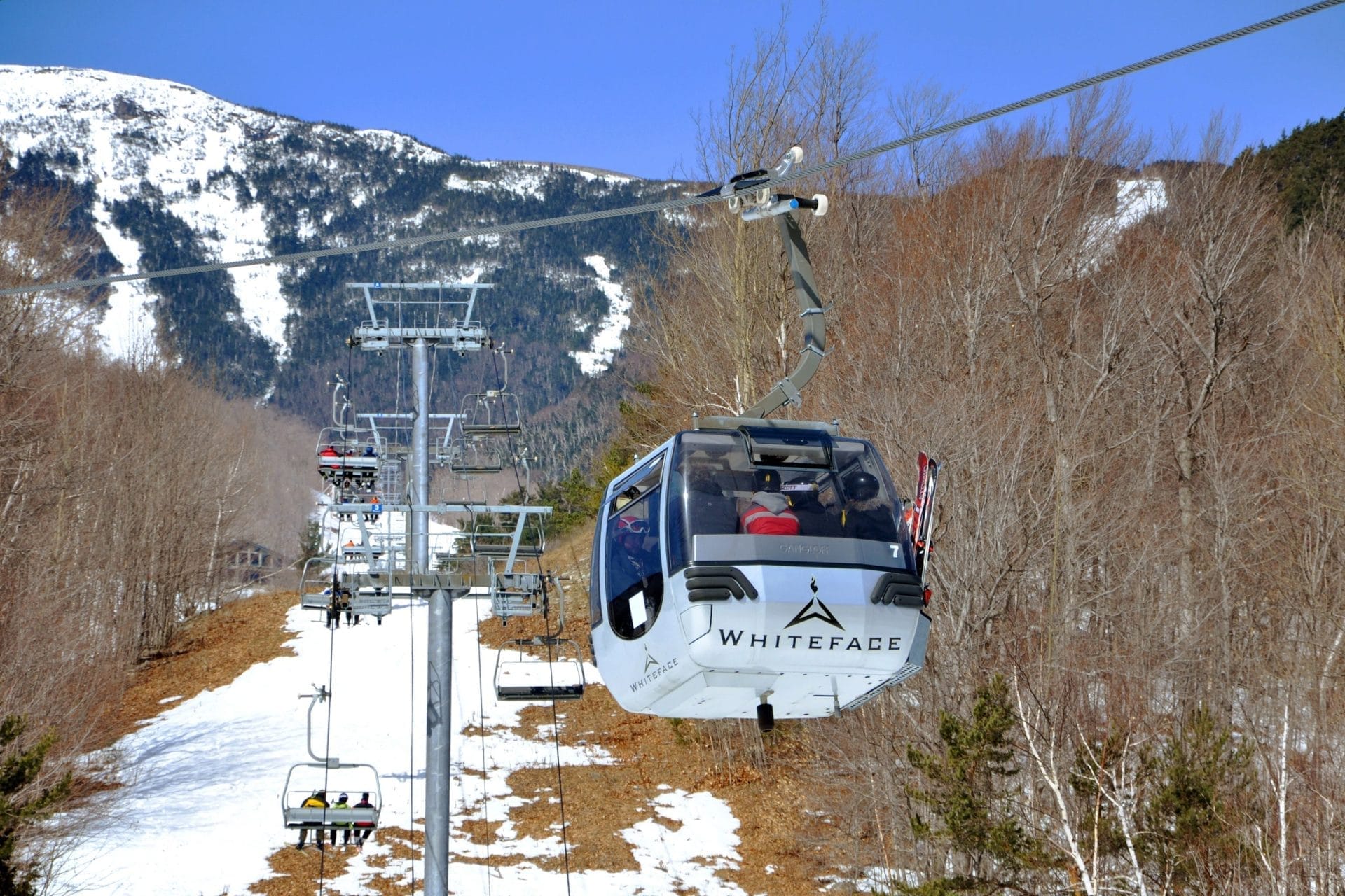 A gondola labeled Whiteface carries eager skiers up a snowy mountain. The ski landscape features bare trees and mountain peaks under a clear blue sky. A gondola labeled Whiteface carries eager skiers up a snowy mountain. The ski landscape features bare trees and mountain peaks under a clear blue sky.