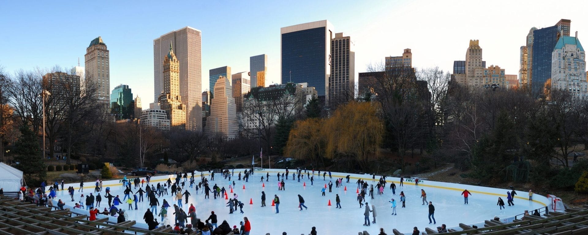 A large group of people ice skating at an outdoor rink in a park, set against the iconic NYC skyscrapers and leafless trees, indicating winter. The clear sky enhances the lively and bustling atmosphere. A large group of people ice skating at an outdoor rink in a park, set against the iconic NYC skyscrapers and leafless trees, indicating winter. The clear sky enhances the lively and bustling atmosphere.