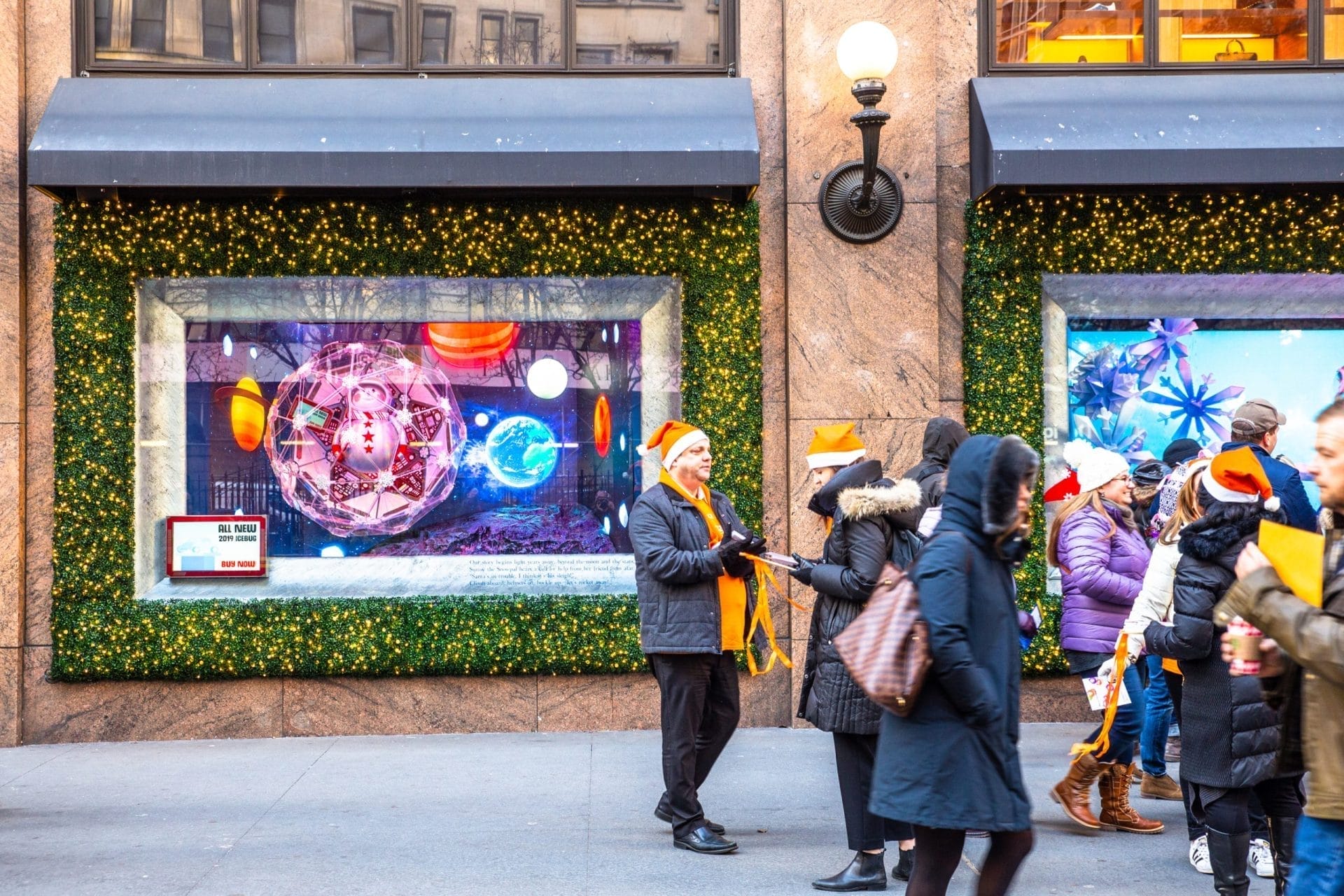 People in winter clothing, some wearing orange Santa hats, gather in front of festive store windows in NYC adorned with green garlands and lights. The windows display colorful holiday decorations, creating a lively and cheerful scene. People in winter clothing, some wearing orange Santa hats, gather in front of festive store windows in NYC adorned with green garlands and lights. The windows display colorful holiday decorations, creating a lively and cheerful scene.
