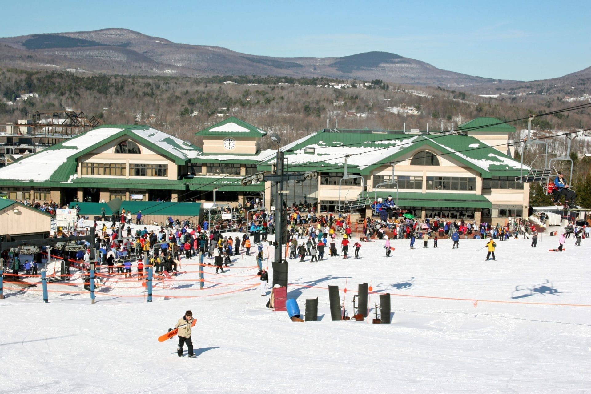 A bustling ski resort features people skiing down a snowy slope, some holding skis and snowboards. A large lodge with green roofs stands against scenic mountains under a clear blue sky. Ski enthusiasts are also using the lift on the right. A bustling ski resort features people skiing down a snowy slope, some holding skis and snowboards. A large lodge with green roofs stands against scenic mountains under a clear blue sky. Ski enthusiasts are also using the lift on the right.