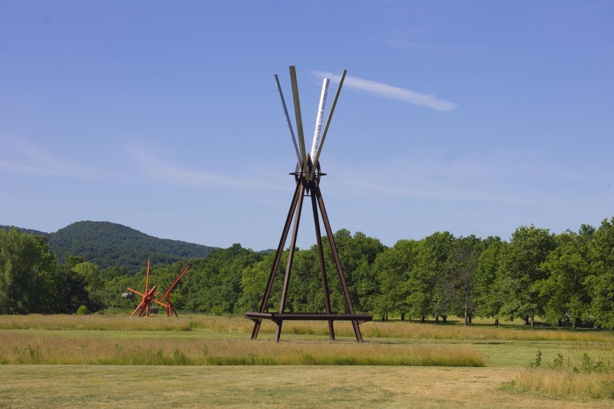A large outdoor sculpture made of long, vertical metal beams arranged in a fan-like structure stands on a grassy field, with another abstract red sculpture in the background. This serene landscape, adorned with trees and hills, makes it one of New York Sculpture Parks worth visiting. A large outdoor sculpture made of long, vertical metal beams arranged in a fan-like structure stands on a grassy field, with another abstract red sculpture in the background. This serene landscape, adorned with trees and hills, makes it one of New York Sculpture Parks worth visiting.