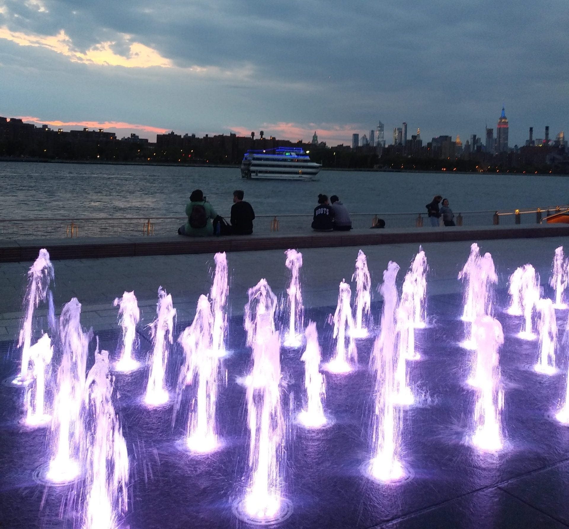 A view of illuminated fountains with purple lights in the foreground, set against a river backdrop, offers one of the off-beat experiences to enjoy in New York City. People sit by the water as a lit-up boat sails by, and the city skyline is visible under a clouded sky with a hint of sunset. A view of illuminated fountains with purple lights in the foreground, set against a river backdrop, offers one of the off-beat experiences to enjoy in New York City. People sit by the water as a lit-up boat sails by, and the city skyline is visible under a clouded sky with a hint of sunset.