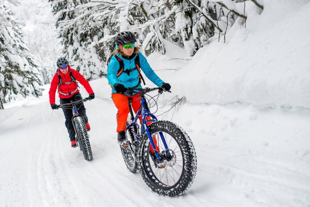 Two people enjoy winter fun in New York State, riding fat bikes on a snowy trail through the forest. Clad in colorful winter gear with helmets and goggles, they pedal past snow-covered trees, immersing themselves in a scenic winter landscape. Two people enjoy winter fun in New York State, riding fat bikes on a snowy trail through the forest. Clad in colorful winter gear with helmets and goggles, they pedal past snow-covered trees, immersing themselves in a scenic winter landscape.