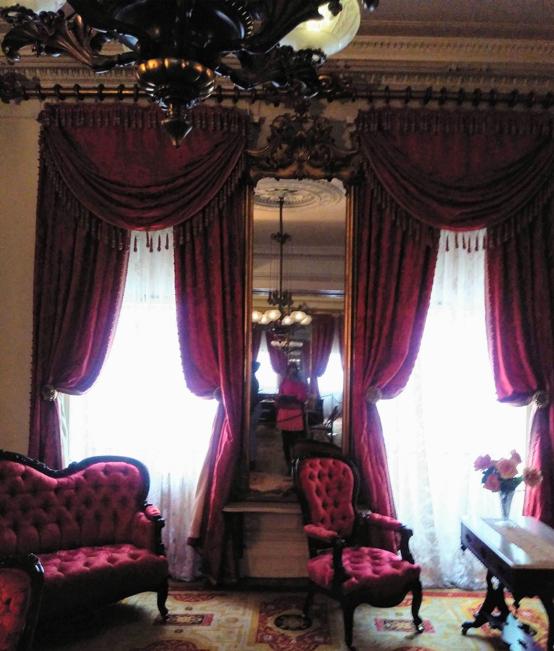 A vintage living room with ornate red velvet curtains and Victorian-style furniture, including a tufted sofa and armchair. The large mirror reflects the chandelier and interior, reminiscent of off-beat experiences to enjoy in New York City. A small table with a vase of flowers completes the setting. A vintage living room with ornate red velvet curtains and Victorian-style furniture, including a tufted sofa and armchair. The large mirror reflects the chandelier and interior, reminiscent of off-beat experiences to enjoy in New York City. A small table with a vase of flowers completes the setting.