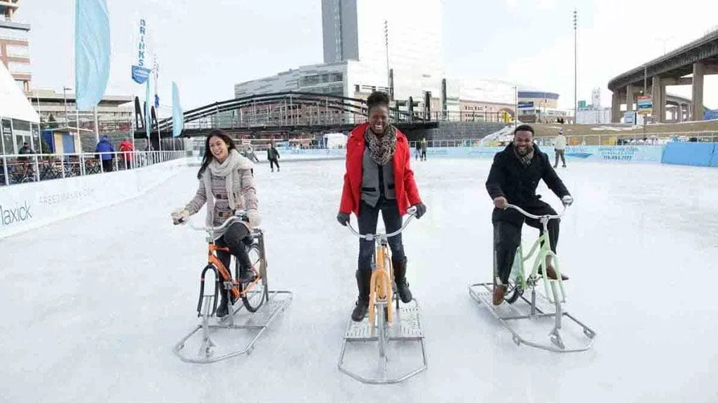 Three people enjoying winter fun in New York State ride ice bikes on an outdoor rink. Theyre smiling and wrapped in cozy winter clothing. Buildings and a highway overpass form the backdrop on this clear, crisp day. Three people enjoying winter fun in New York State ride ice bikes on an outdoor rink. Theyre smiling and wrapped in cozy winter clothing. Buildings and a highway overpass form the backdrop on this clear, crisp day.