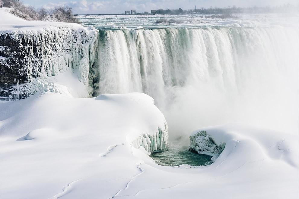 Experience winter fun in New York State with a breathtaking scene of a large waterfall, partially frozen and draped in snow. The water cascades powerfully over the edge, mist rising under a pale blue sky as snow blankets the enchanting landscape. Experience winter fun in New York State with a breathtaking scene of a large waterfall, partially frozen and draped in snow. The water cascades powerfully over the edge, mist rising under a pale blue sky as snow blankets the enchanting landscape.