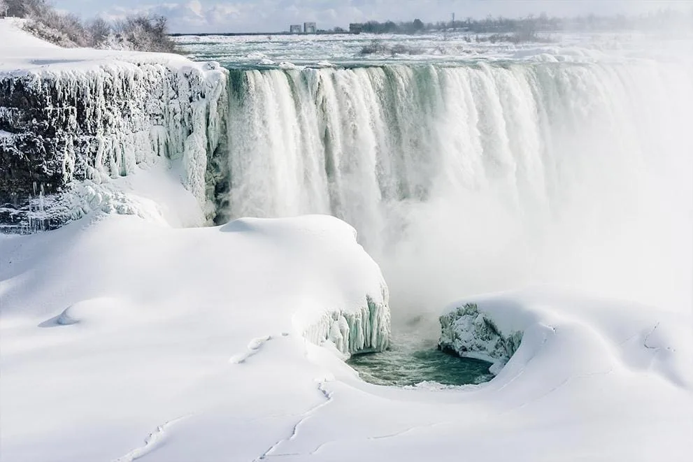 Experience winter fun in New York State with a breathtaking scene of a large waterfall, partially frozen and draped in snow. The water cascades powerfully over the edge, mist rising under a pale blue sky as snow blankets the enchanting landscape. Experience winter fun in New York State with a breathtaking scene of a large waterfall, partially frozen and draped in snow. The water cascades powerfully over the edge, mist rising under a pale blue sky as snow blankets the enchanting landscape.